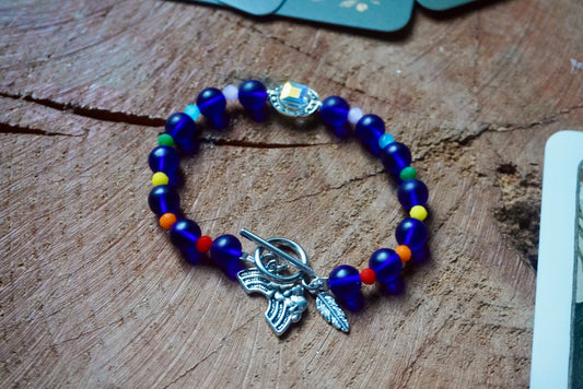 Blue glass beaded bracelet with a silver toned feather charm and rainbow charm. Each blue bead is separated by a different color, in rainbow order. The bracelet is on a wooden surface.
